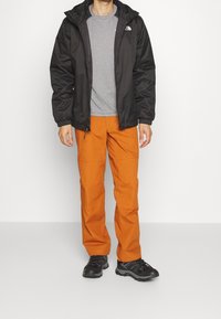 Giacca leggera nera con cappuccio, camicia grigia e pantaloni arancioni. Scarpe da trekking nere con suola testurizzata, il tutto su uno sfondo bianco.