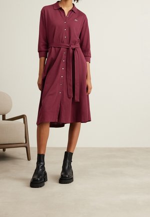 Mujer con un vestido burdeos hasta la rodilla, abotonado, con un cinturón atado a la cintura y botas negras gruesas hasta el tobillo, de pie en el interior junto a una silla beige.