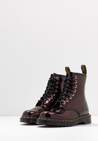 Botas de caña alta con cordones en un material patentado burdeos oscuro, con cordones negros, detalles amarillos y una suela de goma negra gruesa.