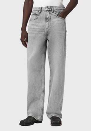 Person iført lysegrå, løstsiddende jeans med hånden i lommen og sorte sko, stående mod en ensfarvet baggrund.