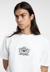 White cotton t-shirt featuring a black embroidered crest with lions and a crown, centered on the chest, short sleeves, casual fit.