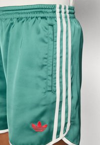 Calções de desporto turquesa feitos de um tecido brilhante, apresentando três riscas brancas de lado e um logótipo vermelho da Adidas na bainha.