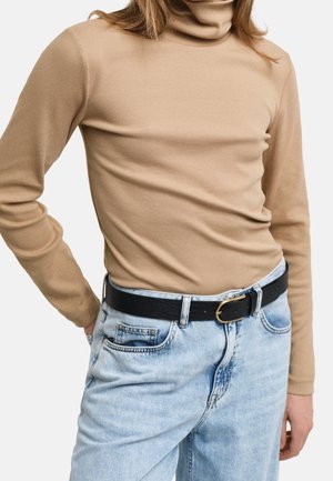 Persona que lleva un suéter de cuello alto beige metido en unos vaqueros azul claro con un cinturón negro con hebilla dorada.