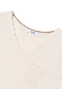 Crèmekleurige top met V-hals, gemaakt van zachte stof, met een klein patchzakje en een geribbelde halsdetail. Etiket leest "CECI."