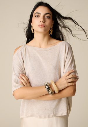 Młoda kobieta z długimi ciemnymi włosami, ubrana w beżowy sweter z wycięciem na ramieniu, kolczyki w kształcie gwiazdek oraz dwie masywne bransoletki, ze skrzyżowanymi ramionami.