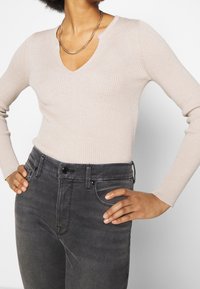 Béžový žebrovaný svetr s dlouhým rukávem a výstřihem do V, kombinovaný se šedými džínami s vysokým pasem. Svetr má jemnou texturu; džíny jsou zdobené černými knoflíky.