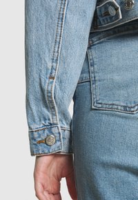 Svetle modrá džínová bunda s rukávem a knopkovým manžetem, ukazující kontrastní šití a hladký povrch. Detailní záběr ruky na látce.