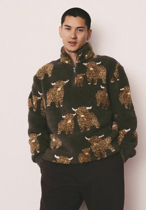Junger Mann in einem dunklen Fleece-Pullover mit rötlichen und weißen Highland-Rind-Mustern, stehend mit Händen in den Taschen vor einem hellen Hintergrund.