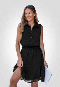 Een vrouw glimlacht terwijl ze een mouwloos zwart jurkje met stippen draagt, een witte clutch vasthoudt en de zoom van het jurkje aanpast.