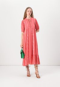Long, pink floral dress with puff sleeves, button details, and a tiered hem. Model holds a small green handbag. Sandals feature ankle ties.