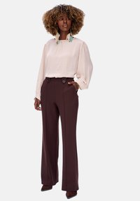 Lichtroze blouse met lange mouwen en een geplooide halslijn, gecombineerd met hooggesneden bruine broek met twee knopen en zijzakken.