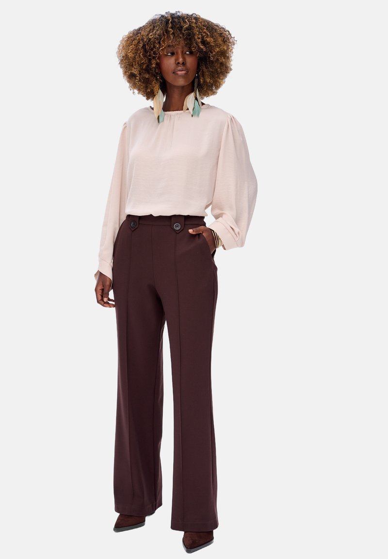 Lichtroze blouse met lange mouwen en een geplooide halslijn, gecombineerd met hooggesneden bruine broek met twee knopen en zijzakken.