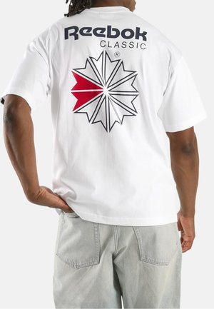 Mann trägt weißes Reebok Classic T-Shirt mit schwarzem und rotem Stern-Logo auf dem Rücken, kombiniert mit hellgrauen Jeans, steht mit der Hand in der Hüfte.