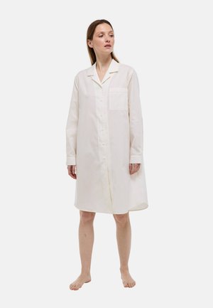 Femme debout pieds nus portant une robe chemise blanche à manches longues, longueur genou, boutonnée avec une poche poitrine, sur un fond uni.