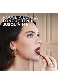 Rouge à lèvres liquide appliqué avec un applicateur. Le modèle a un teint lisse, des cheveux foncés et des boucles d'oreilles en or. Fond neutre.