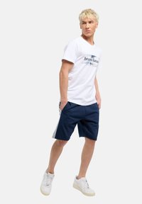 Ung man med blont hår som bär en vit t-shirt med grafik, marinblå shorts med sidosträck, vita sneakers, står med händerna i fickorna.