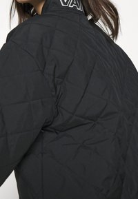 Sort quiltet jakke med høj krave og broderet logo på baghalsen, der fremviser et glat, tekstureret stof.