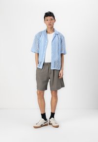 Lyseblå blonder kortermet skjorte, hvit t-skjorte under, grå shorts og hvite joggesko med svarte detaljer; ensfarget bakgrunn.
