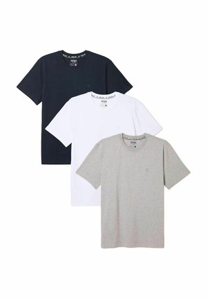 Mann trägt ein hellgraues T-Shirt, hellblaue Jeans und braune Sneakers und steht vor einer weißen Wand.