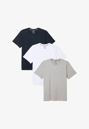 Mann trägt ein hellgraues T-Shirt, hellblaue Jeans und braune Sneakers und steht vor einer weißen Wand.