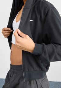 Person som bär en mörk zip-up jacka över en vit crop top och mörka byxor, händerna håller jackan öppen, vilket visar magen med navelpiercing.