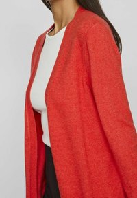 Röd kofta i mjukt stickat material, med öppen framsida och avslappnad passform, bärs över en vit topp. Slät textur med minimal detaljering.