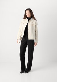 Cremefarbene Jacke mit strukturiertem Material und goldenen Knöpfen, zwei Brusttaschen und ausfransenden Säumen, kombiniert mit schwarzen, hochtaillierten Hosen und spitzen schwarzen Schuhen.