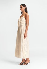 Helles beige Maxikleid mit schwarzen Punkten, gerafftem Oberteil, verstellbaren Spaghetti-Trägern und einem fließenden Rock. Getragen mit schwarzen Flip-Flops.