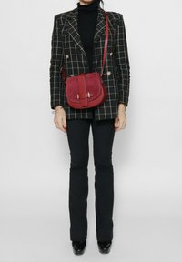 Borsa a tracolla in suede rosso con forma curva, dotata di una cintura centrale e dettagli in metallo. Giacca a quadri bianca e nera sullo sfondo.