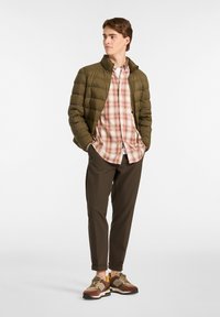 Chaqueta acolchada oliva, camisa a cuadros rosa y blanca, pantalones marrones y zapatillas deportivas marrones y mostaza. Posición de caminata, manos en los bolsillos.