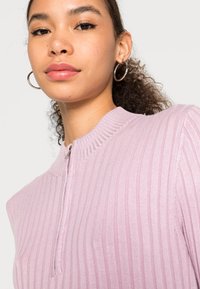 Top de malha canelada lilás claro com gola alta e um pequeno fecho éclair. Apresenta um acabamento texturizado e discretos detalhes brilhantes.