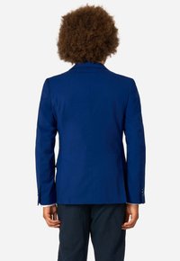 Tumši zils žakets ar piegriezumu, vienu atveri un divu pogu aizdari. Gluda auduma tekstūra, iedobti mēteļa uzliku un pogātu aproču.