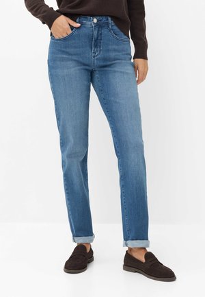 Person, die hellblaue umgeschlagene Jeans und dunkelbraune Loafers trägt, mit einer Hand in der Tasche und einem dunkelbraunen Pullover.
