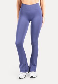 Leggings atletici aderenti viola con fascia alta in vita, realizzati in tessuto liscio; svasati alle caviglie, abbinati a scarpe da ginnastica bianche.