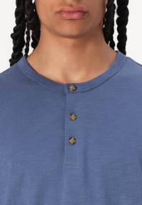 Blå kortermet henley skjorte med rund hals og tre brune knapper ved placket. Stoffet har en teksturert overflate.