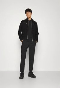 Črna jakna z zadrgo čez črno majico, kombinirana s črnimi hlačami in črnimi supergami. Gladek material, minimalističen dizajn in prilagojena oblika.