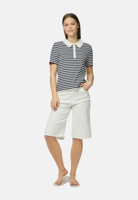 Jūras zila un balta svītrainā polo krekls ar baltu apkakli, savienots ar baltām ceļgala garuma šortiem. Modelis valkā gaišas čības un stāv vienkārši.