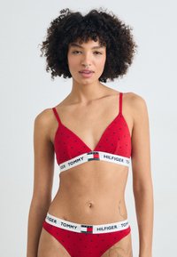 Crvena pamučna grudnjak s tankim trakama i trokutastim šalicama, s malim crnim uzorcima srca. Traka ima plavo-bijeli "TOMMY HILFIGER" logotip.