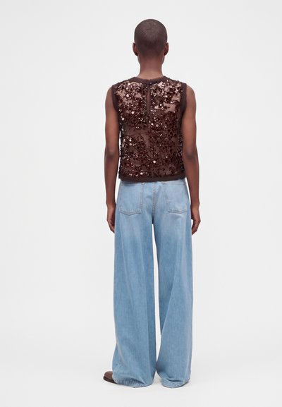 Personne portant un haut sans manches marron à sequins et un jean large bleu clair, debout dos à un fond uni.