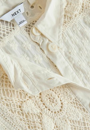 Kreemmikas pluus keeruka tikandi, pitsdetailide ja pärlmutter nööbidega. Omadused: kraega disain ja kergest materjalist.