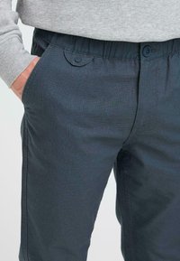 Mørkeblå bukser med en glat tekstur, elastisk talje og knapdetalje på lommerne. Stoffet har et subtilt mønster.