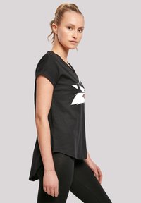 Svart t-skjorte med hvit grafisk design på forsiden, korte ermer og en litt lengre bakhem. Modell som bruker svarte leggings.