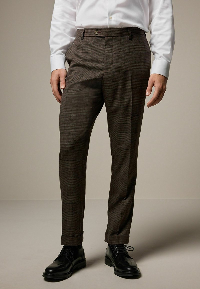 Homme portant un pantalon habillé à carreaux marron foncé, une chemise blanche et des chaussures en cuir noires, debout devant un fond uni.