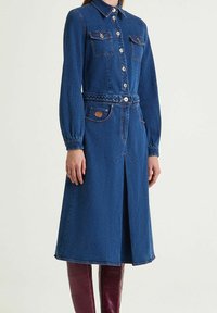 Robe-chemise en denim bleu foncé avec manches longues, fermeture boutonnée à l'avant, deux poches poitrine et ceinture tressée. Dotée d'une fente à l'avant.