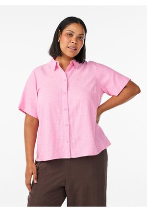 Femme aux cheveux bruns mi-longs, portant une chemise rose à manches courtes et un pantalon marron foncé, posant avec une main sur la hanche.