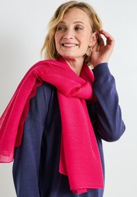 Bufanda de color rosa intenso con textura acanalada, caída con gracia, llevada sobre un suéter de color azul marino. Tejido suave con acentos brillantes. Sin patrones ni herrajes.