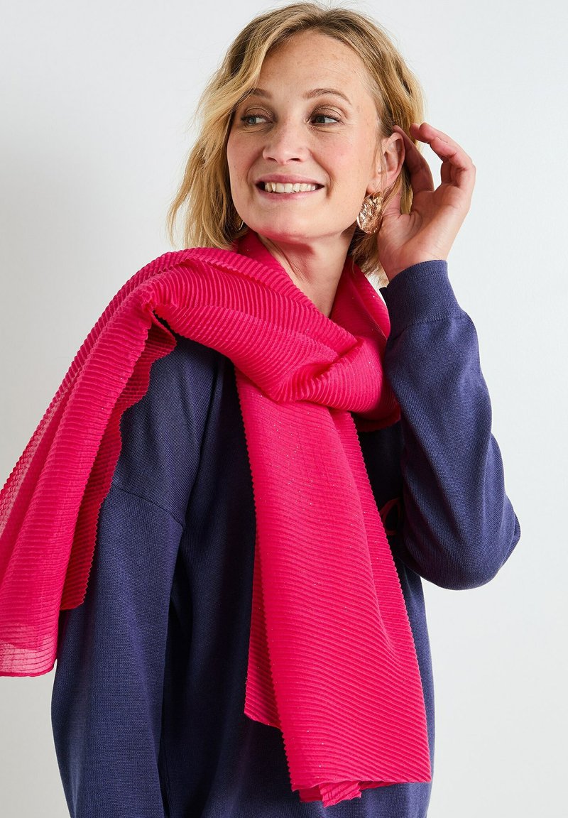 Bufanda de color rosa intenso con textura acanalada, caída con gracia, llevada sobre un suéter de color azul marino. Tejido suave con acentos brillantes. Sin patrones ni herrajes.