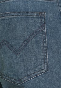 Detailní záběr na modrou džínovou látku s zadní kapsou s cik-cak prošitím v tmavě modré barvě. Textura vykazuje jemné variace v odstínech.