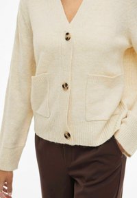 Bež kardigan s gumbima i dva prednja džepa, nosi se preko tamnosmešnih hlača, ruka opušteno odmarajući u džepu.