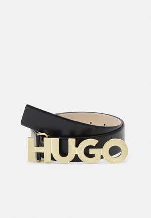 Ceinture en cuir noir enroulée avec une grande boucle dorée formant les lettres HUGO sur un fond clair uni.